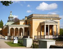 Historisches, klassizistisches Gebäude mit gelber Fassade und Säulenportalen, flankiert von einem überdachten Arkadengang. Im Hintergrund erhebt sich ein Turm mit einer Uhr und einer vergoldeten Skulptur auf der Spitze.