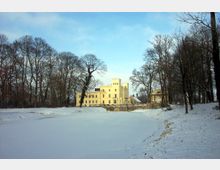 Gelbes Schloss im neoklassizistischen Stil, umgeben von kahlen, winterlichen Bäumen und einer schneebedeckten Landschaft, im Vordergrund ein zugefrorener See. Im Hintergrund heller Himmel mit vereinzelten Wolken.