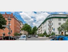 Straßenszene in einem Wohngebiet mit mehrstöckigen Wohngebäuden; links ein orangefarbenes Gebäude, rechts ein weißes mit grünen Dächern und Blumen an den Balkonen. Die Straße ist von geparkten Autos gesäumt, und Bäume stehen in der Mitte der Fahrbahn entlang eines Gehwegs; Straßenschilder sind deutlich sichtbar.