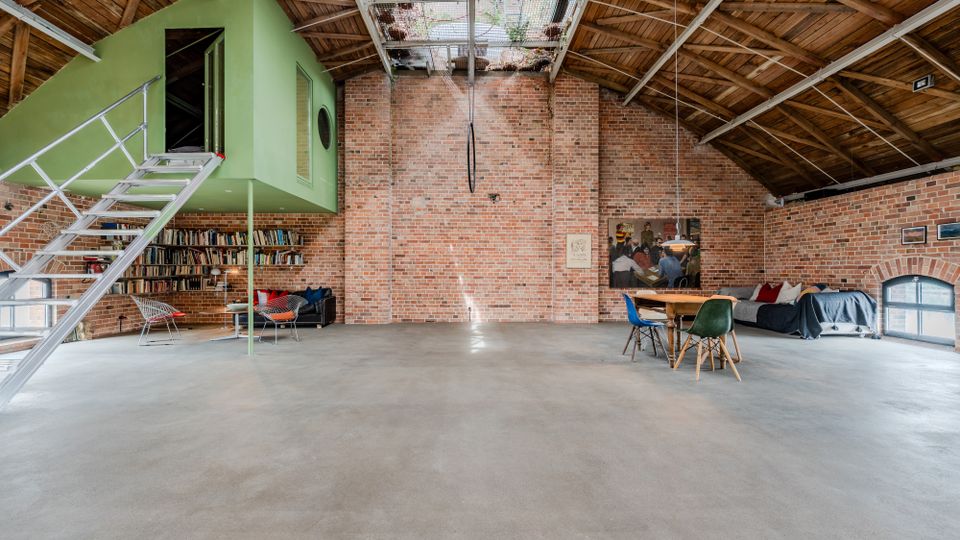 Großer Loft- oder Atelierraum mit Backsteinwänden, sichtbarem Holzdachstuhl und einem zentralen Oberlicht. Links hängt ein grünes Kubus-Zimmer mit Metalltreppe über einem Bücherregal, rechts stehen ein Esstisch mit Stühlen und ein Sofa vor einem bogenförmigen Fenster.