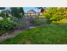 Gartenbereich mit Rasenfläche im Vordergrund und einem Blumenbeet mit Lavendel, Stauden und kleinen Solarlampen entlang eines schmalen Wegs. Im Hintergrund sind ein Zaun, hohe immergrüne Sträucher sowie ein Wohnhaus und ein kleiner Holzunterstand zu sehen.