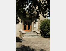 Eingang eines historischen Gebäudes mit steinernen Stufen, hölzernen Doppeltüren mit metallenen Verzierungen und Laternen auf beiden Seiten. Der Vorplatz ist mit Pflastersteinen bedeckt, im Vordergrund hängen Blätter eines Baumes.