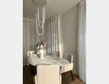 Heller Essbereich mit einem ovalen weißen Tisch, umgeben von beige gepolsterten Stühlen. Eine minimalistische hängende Leuchte und ein Blumenstrauß in einer weißen Vase auf dem Tisch ergänzen den Raum, der von dünnen weißen Vorhängen und natürlichem Licht geprägt ist.