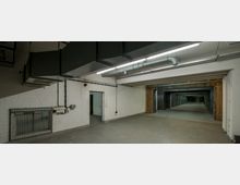 Leerer Raum mit Betonboden, weißen Ziegelwänden und sichtbaren Belüftungssystemen an der Decke. Im Hintergrund führen mehrere Metallschleusen und Holzrahmen in eine andere, tiefer liegende Räumlichkeit.