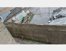 Eingezäuntes, verlassenes Gelände mit zerfallenden Mauerresten, Trümmern und einer Betonfläche, teilweise bedeckt mit Pfützen. Im Hintergrund liegen Schutthaufen unter grauen Planen.