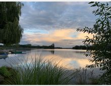 Ein See in einer natürlichen Umgebung bei Sonnenuntergang, umgeben von Bäumen und Schilf am Ufer. Im Vordergrund sind Gräser und ein Weidenbaum, während am Ufer kleine Anlegeboote liegen.