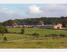 Ein ländlicher Hof mit roten Backsteingebäuden und dunkelgrauen Dächern, umgeben von weitläufigen, grünen Wiesen und Holzzäunen. Im Hintergrund ist ein Wald zu sehen, der teilweise von herbstlich gefärbten Bäumen durchzogen ist.
