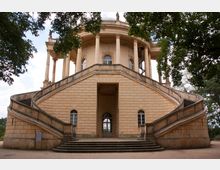 Gelblich-beiges, klassizistisches Rundgebäude mit geschwungenen Doppeltreppen, hohen Säulen und rundbogenförmigen Fenstern, umgeben von Bäumen. Eine offene Plattform mit Balustraden und Statuen ist auf der oberen Ebene erkennbar.