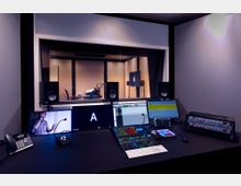 Tonstudio mit einem modernen Regie-Arbeitsplatz, ausgestattet mit mehreren Monitoren, einem Mischpult, Lautsprechern und Audiogeräten. Durch ein großes Studiofenster ist der schallisolierte Aufnahmebereich mit Mikrofonen und einem Bildschirm sichtbar.