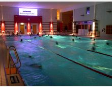 Hallenbad mit beleuchtetem Schwimmbecken und mehreren Schwimmbahnen, umgeben von Fliesenboden. Im Hintergrund sind Umkleidekabinen, Türen, und ein Schild mit der Aufschrift "Solarent Bräunungsstudio" zu sehen.