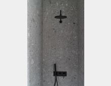 Duschkabine mit grauen Terrazzo-Wänden, einer schwarzen Regenduschkopfarmatur oben und einer schwarzen Handbrause sowie Bedienelementen an der Wand.