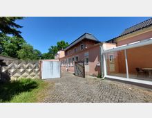 Ein rosa verputztes Gebäude mit Satteldach und mehreren weißen Fenstern, daneben eine gläserne Veranda. Im Vordergrund liegt ein gepflasterter Hof mit einem Metalltor, begrenzt durch eine Betonmauer; im Hintergrund sind Bäume und blauer Himmel sichtbar.