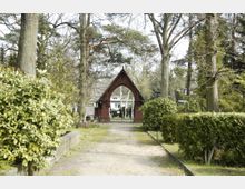 Sandweg führt durch einen grünen Park mit Bäumen und Büschen zu einem roten, spitzbogigen Backsteintor mit einem schmiedeeisernen Gitter. Dahinter sind Wohnhäuser sichtbar.
