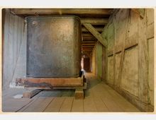 Holzrahmenkonstruktion im Inneren eines historischen Gebäudes mit sichtbaren Balken und Fachwerk. Im Vordergrund steht ein massiver, metallener Behälter auf einer Holzplattform, der Raum ist von Holzböden und rauen Wänden geprägt.