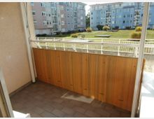 Blick von einem Balkon mit Fliesenboden und einer Sichtschutzwand aus Bambus auf eine grüne Rasenfläche mit dekorativen Gräsern. Im Hintergrund sind mehrstöckige Wohngebäude in hellen Farben zu sehen.