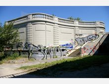 Historische Betonmauer mit Balustrade, teilweise von Graffitis bedeckt, an der sich eine Treppe entlangzieht. Im Vordergrund ist eine grasbewachsene Fläche und ein gepflasterter Weg zu sehen, dahinter ein klarer blauer Himmel.