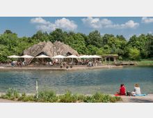 Eine Cafeteria mit einer felsartigen, organisch geformten Architektur steht am Rand eines kleinen Sees, umgeben von üppiger Vegetation. Menschen sitzen unter weißen Sonnenschirmen oder spazieren am Ufer; im Vordergrund sitzen zwei Personen auf einer Wiese am Wasser.
