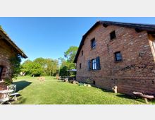Backsteingebäude mit schwarzen Fensterläden auf einem weitläufigen, grünen Rasen. Im Hintergrund sind Bäume und Gartenmöbel unter einem Sonnenschirm zu sehen, bei klarem Himmel.