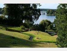 Ein Park mit leicht hügeliger Landschaft, einem kleinen Teich und einem geschwungenen Pfad im Vordergrund. Im Hintergrund sind Bäume, ein Fluss und eine Stahlbrücke sichtbar.
