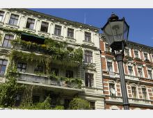 Alttext: Mehrstöckiges historisches Wohngebäude mit verzierten Fassaden und zahlreichen Fenstern, von denen einige mit Blumen bepflanzt sind. Ein Balkon ist stark mit Pflanzen überwuchert, und im Vordergrund steht eine Straßenlaterne aus Metall vor einem blauen Himmel.