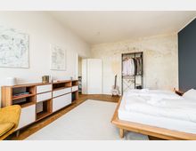 Helles Schlafzimmer mit Holzbett und weißer Bettwäsche auf Fischgrätparkett, dazu ein langer Holz-Sideboard mit offenen Fächern und abstrakten Bildern an der Wand. An der Rückwand stehen ein offenes Kleiderregal und eine Gitarre vor einer teils roh wirkenden, hellen Wandfläche.