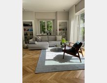 Helles Wohnzimmer mit grauem Ecksofa, einem runden Glastisch und einem Polstersessel mit Holzrahmen. Der Raum hat einen hölzernen Fischgrätenparkettboden, ein großes Fenster mit weißen Vorhängen und Regale mit farbiger Dekoration an den Wänden.