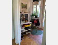 Flur einer Wohnung mit Garderobe und Stauraum. Zu sehen sind ein Schuhregal mit mehreren Paar Schuhen, eine Pinnwand mit kleinen Ablagekörben, eine Garderobe mit Jacken, ein grüner Stuhl auf einem runden Teppich und ein großes Fenster, das den Raum erhellt.