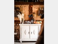 Rustikale Kaffeestation mit einer weißen, verzierten Kommode vor einer Holzwand mit Lehmstruktur. Auf der Kommode stehen Gläser, eine Kaffeemaschine und Trockenblumen, daneben hängt eine kleine Tafel mit der Aufschrift „Coffee Bar“.