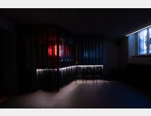 Ein dunkler Innenraum mit moderner Bar, gekennzeichnet durch vertikale schwarze Metallstreben und roten sowie blauen Lichteffekten. Im Vordergrund stehen mehrere Hocker, seitlich befindet sich ein teilweise beleuchtetes Fenster.