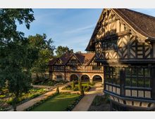 Historisches Fachwerkgebäude mit roten Ziegeldächern, großen, verzierten Giebelfronten und Spitzbogenarkaden. Im Vordergrund ein gepflegter Garten mit Rasenfläche, Blumenbeeten und geschnittenen Hecken, flankiert von hohen Bäumen.