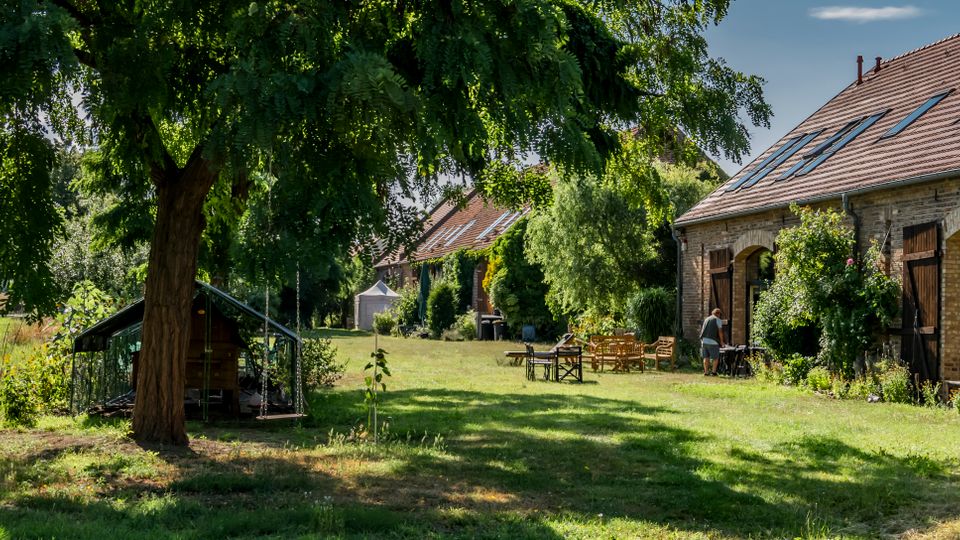 Ländlicher Hofgarten mit großer Rasenfläche, einem schattenspendenden Baum und einem kleinen Gewächshaus am Rand. Rechts stehen umgebaute Backsteingebäude mit Ziegeldach, Dachfenstern und großen Holztoren, davor Gartenmöbel und üppige Bepflanzung.