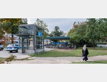 Eingang zur U-Bahn-Station "Amrumer Straße" in Berlin mit einer gläsernen Aufzugsanlage und einem überdachten Fahrradstellplatz im Hintergrund. Umgeben von Bäumen und Grünflächen, führt ein Weg durch den Parkbereich.