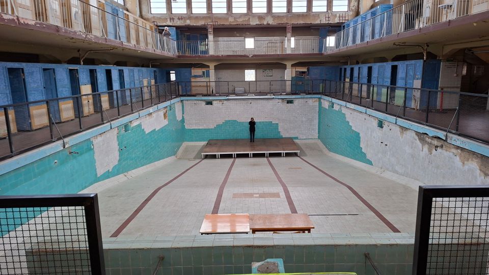 Ehemaliges, leer stehendes Schwimmbad mit türkisen Fliesen, abgenutzten Wänden und einem leeren Becken. Zwei Galerien mit metallenen Geländern und blauen Umkleidekabinen umrahmen den Raum, und Licht fällt durch große Fenster im oberen Bereich.