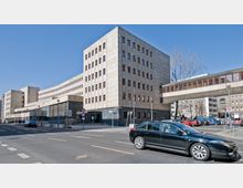 Ein großes, modernes Verwaltungsgebäude mit einer Fassade aus beige-grauen Kacheln und zahlreichen Fenstern. Eine verglaste Fußgängerbrücke verbindet zwei Gebäudeteile über eine Straße, auf der Autos parken und fahren; im Hintergrund sind weitere Gebäude sichtbar.