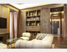 Luxuriöses Wohnzimmer mit Holzvertäfelung, eingebautem Regal mit Büchern und Dekorationen, gepolstertem Sofa, rundem Glastisch mit Metallgestell und Plüschhocker. Im Hintergrund führt eine Tür zu einem Flur mit modernem Design.