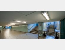 Unterirdischer Fußgängertunnel mit gefliesten Wänden in Hellgrün, beleuchtet durch Deckenleuchten. Treppen und eine Rolltreppe führen nach oben, links ist ein breiter Durchgang sichtbar.