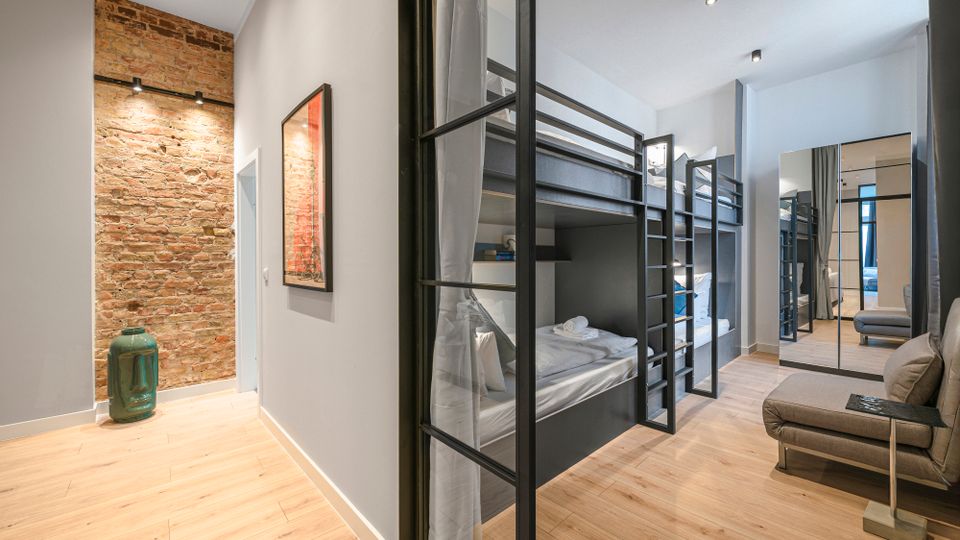 Moderner Schlafsaal im Hostelstil mit mehreren schwarzen Etagenbetten und Vorhängen, hellen Holzböden und weißen Wänden. Links ist eine freiliegende Ziegelwand mit Wandlampen, am Ende ein Spiegel und rechts ein kleines Sitzmöbel zu sehen.