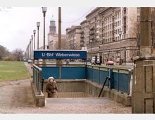 Eingang zur U-Bahn-Station Weberwiese in Berlin, mit einem blauen Schild und Treppenabgang, umgeben von einem breiten Gehweg. Im Hintergrund sind Wohngebäude im sowjetischen Stil, Straßenlaternen und eine Kirchturmspitze zu sehen, daneben eine grüne Rasenfläche.