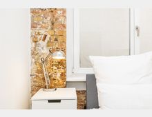 Schlafzimmer-Ecke mit einem Bett mit weißen Kissen neben einem weißen Nachttisch und einer verstellbaren Metall-Schreibtischlampe. Dahinter sind eine freigelegte Ziegelwand und ein großes Fenster mit weißem Rahmen zu sehen.