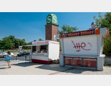 Im Vordergrund stehen zwei Imbissstände, einer für Quarkkeulchen mit geöffnetem Verkauf und einer mit geschlossenem Rollladen, daneben eine markante Backsteinsäule mit grünem „S“-Zeichen. Im Hintergrund sind Bäume und ein Straßenbereich mit parkenden und fahrenden Autos sichtbar.