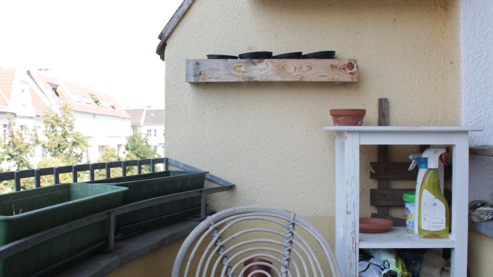 Small apartment balcony with a wooden table and white metal chair beside a railing lined with empty planter boxes. A pale stucco wall has rustic wooden shelves and a small white storage cabinet holding pots and gardening supplies, with neighboring rooftops visible in the background.