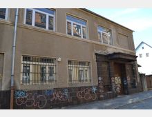 Zweistöckiges Gebäude mit beigen Wänden und einem Eingangsbereich aus dunklen Fliesen, an dem Graffiti angebracht sind. Die Fenster im Erdgeschoss sind vergittert, und auf dem Bürgersteig vor dem Gebäude sind Pflastersteine zu erkennen.