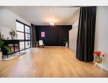 Ein minimalistischer Innenraum mit hellem Holzboden, einer schwarzen Wand mit Vorhängen, einem großen Fenster auf der linken Seite und mehreren Pflanzen als Dekoration. Auf der rechten Seite stehen weiße Einbauschränke, und ein Kunstposter sowie ein Schaukelstuhl akzentuieren den Raum.