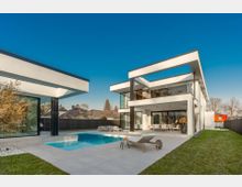 Moderner, zweigeschossiger Flachdachbau mit großen Glasfronten und umlaufendem Balkon, der sich zu einer gepflasterten Terrasse mit rechteckigem Swimmingpool öffnet. Der Außenbereich ist von Rasenflächen und einem hohen Zaun umgeben, im Hintergrund sind Nachbarhäuser und Bäume zu sehen.