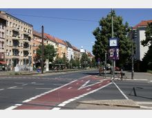 Straßenkreuzung in einer städtischen Umgebung mit mehrstöckigen Wohnhäusern im Hintergrund, die teilweise pastellfarben gestrichen sind. Im Vordergrund sind Fahrspuren mit markierten Radwegen und ein Fahrradfahrer, daneben eine mit Bäumen gesäumte Straße und eine Uhr auf einem Mast.
