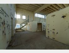 Industriehalle in einem Zustand der Verwitterung mit bröckelnden Wänden, sichtbaren Beschädigungen und abblätternder Farbe. Eine Metalltreppe führt zu einer erhöhten Plattform, Fenster mit einfachem Rahmen lassen Tageslicht einfallen.