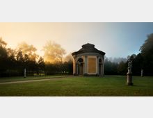 Ein Rundpavillon mit gelber Fassade und Säulen steht auf einer offenen Rasenfläche, umgeben von Bäumen und vereinzelten Statuen. Im Hintergrund erzeugt aufsteigender Nebel und das warme Licht der aufgehenden Sonne eine stimmungsvolle Atmosphäre.
