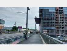 Fußgängerweg auf einer Brücke neben einer stark befahrenen Straße, rechts ein großes Gebäude in Bau mit Gerüsten, links Werbetafeln, Bäume und weitere Stadtgebäude. Der Himmel ist leicht bewölkt.