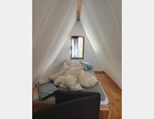 Kleines Schlafzimmer im Dachboden mit stark geneigten, weiß verkleideten Wänden und sichtbarem Holzbalken am First. Ein niedriges Bett steht auf Holzdielen vor einem kleinen Fenster mit Holzrahmen und Vorhang.