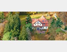 Luftaufnahme eines weißen Landhauses mit rotem Satteldach, umgeben von dichtem Wald und einer Wiese, die mit herbstlichem Laub bedeckt ist. Vor dem Haus befindet sich eine Terrasse mit Tischen und Bänken.